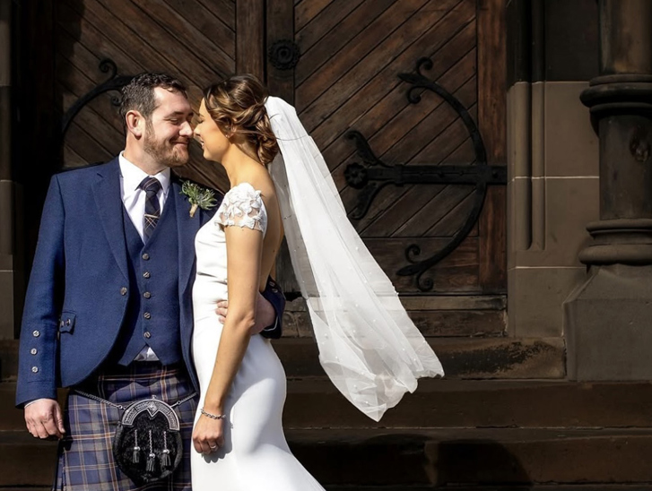 Bride wearing veil and groom wearing a kilt standing outside venue in an embrace while smiling at each other